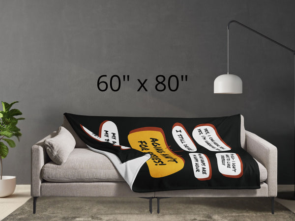 Lying across a grey couch is a large black throw blanket with a large yellow quote bubble in the center that says, Aging Aint For Sissies, Several quotes surround the center quote with several funny old people quips. Size is 60 by 80 inches.