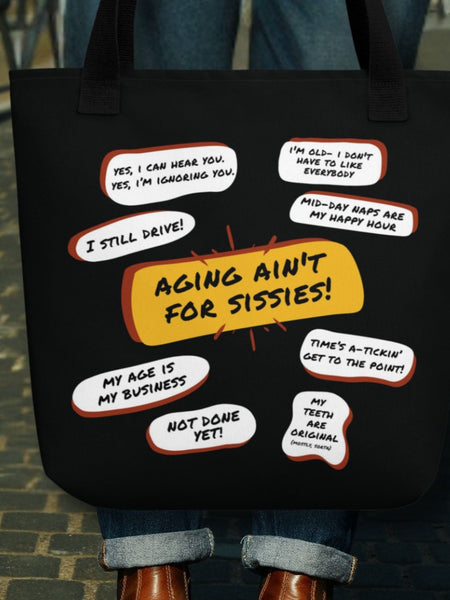 Woman holding a  a black tote bag with several funny old people bubble quotes, including, Aging Ain't For Sissies, I Still Drive, Not Done Yet, and several more.