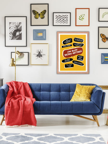 On a gallery wall above a blue couch is a large yellow poster print with a large red quote bubble in the center that says, Im Busy Being Retired. Several move quotes surround it with funny retirement quips. The print is in a natural wood frame.