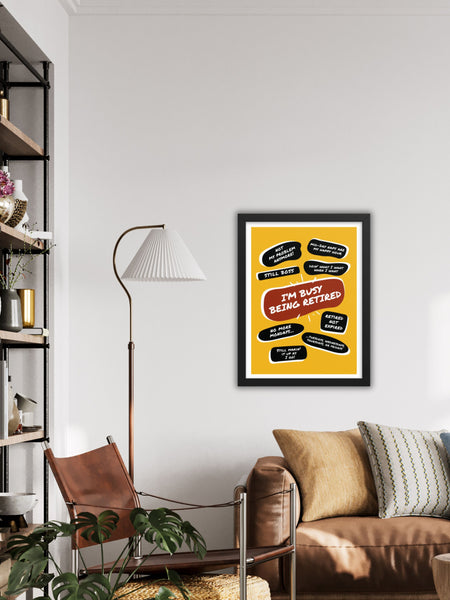 On a wall above a leather couch is a large yellow poster print with a large red quote bubble in the center that says, Im Busy Being Retired. Several black quotes surround the center quote with funny retirement quips. The print is in a black frame.