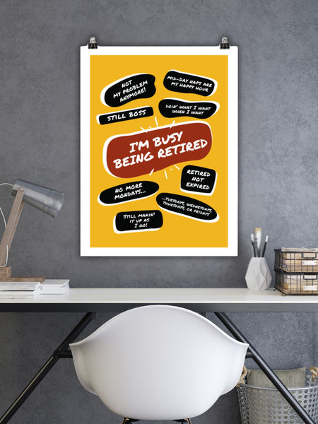 On a grey wall above a white desk is a large yellow poster print with large red quote bubble in the center that says, Im Busy Being Retired. Several quotes surround it with funny retirement quips such as Still Boss, No More Mondays, and many more.