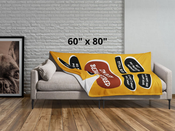 Lying across a grey couch is a large yellow throw blanket with a large red quote bubble in the center that says, I'm Busy Being Tired. Several black quote bubbles surround the center quote with several funny old people quips. Size 60 by 80 inches.