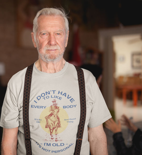 Image of an old man with white hair and beard wearing suspenders and a cream colored shirt that says, "I don't have to like everybody, I'm old, it's not personal" with an red illustration of a cowboy on a horse.