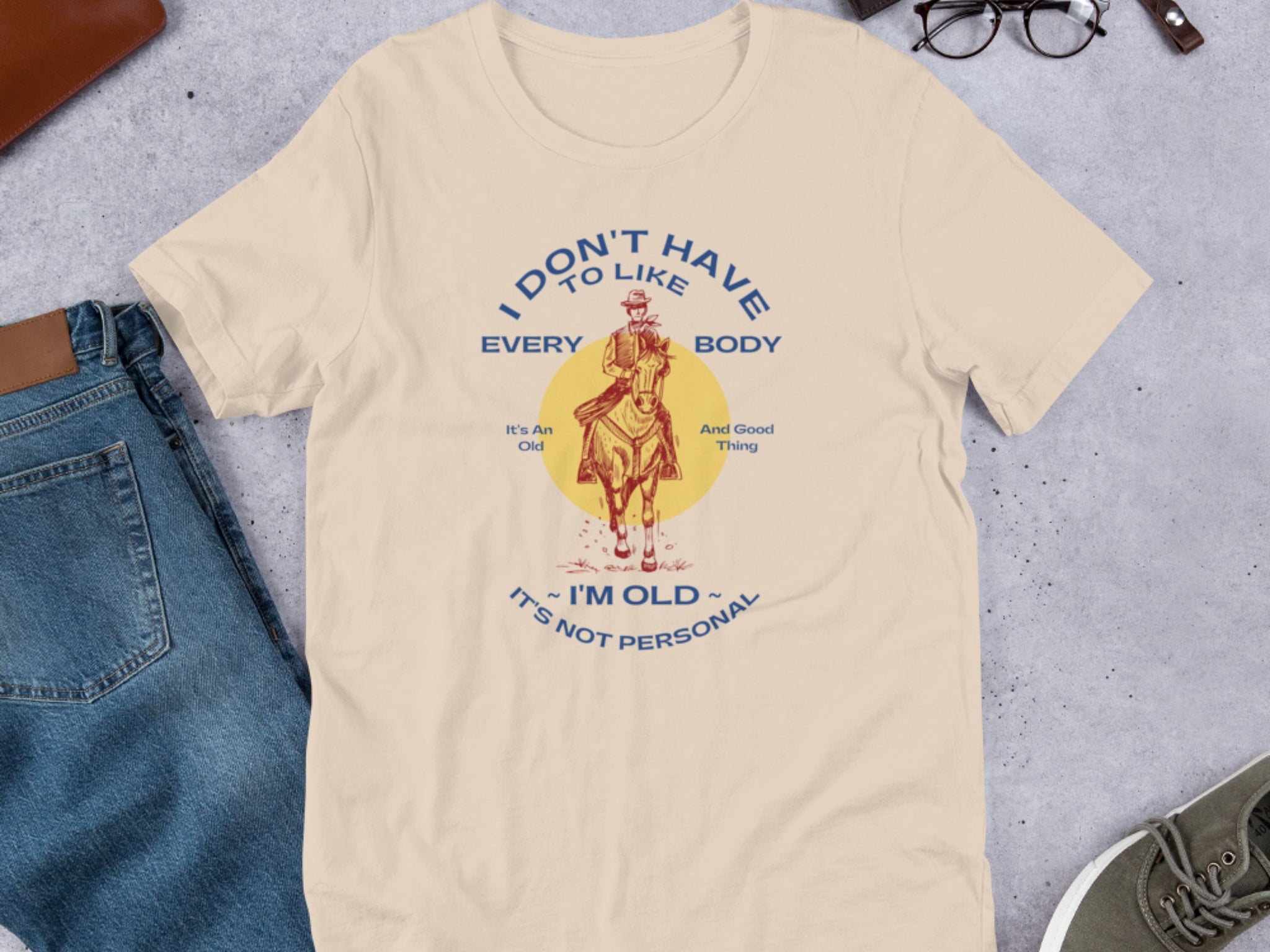 Image of a cream colored shirt that says, "I don't have to like everybody, I'm old, it's not personal" with a vintage red illustration of a cowboy on a horse, surrounded by jeans, glasses, and sneakers.