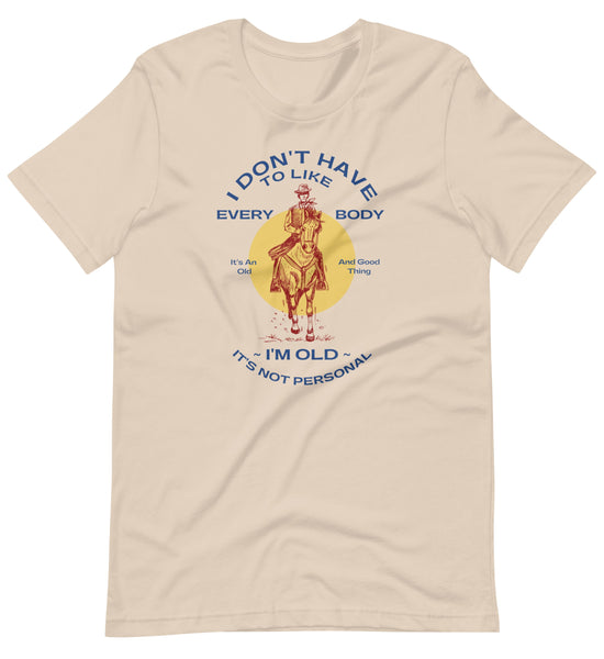 Image of a cream colored shirt that says, "I don't have to like everybody, I'm old, it's not personal" with a vintage red illustration of a cowboy on a horse.