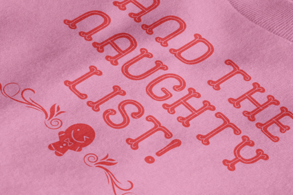 Close up detail of a flat lay mockup of the back of a pink tshirt with text on the outside label  that says, And the naughty list. A small winking gingerbread graphic is below the text.